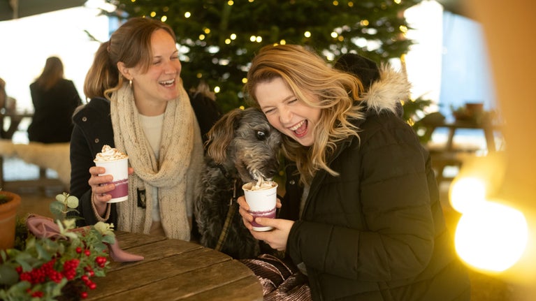 Two women laugh as a dog licks the cream from one of their hot chocolates under Saltram's Christmas tepee